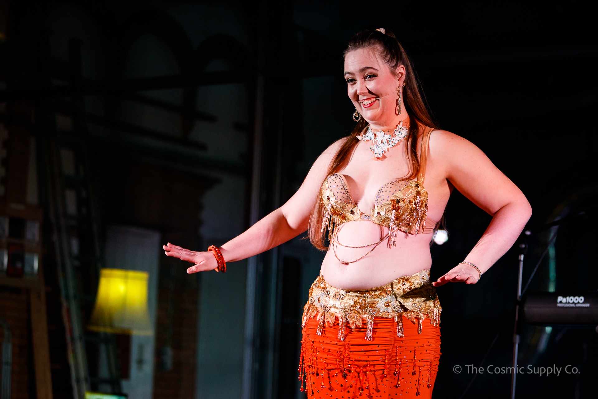 Amanda Salma optræder til 'Mavedans og Koncert' på Ewii House i Kolding. Hun har orange kostume på og holder en Madonna-mikrofon, mens hun instruerer publikum i mavedans. Mørk baggrund fremhæver hende som fokus.